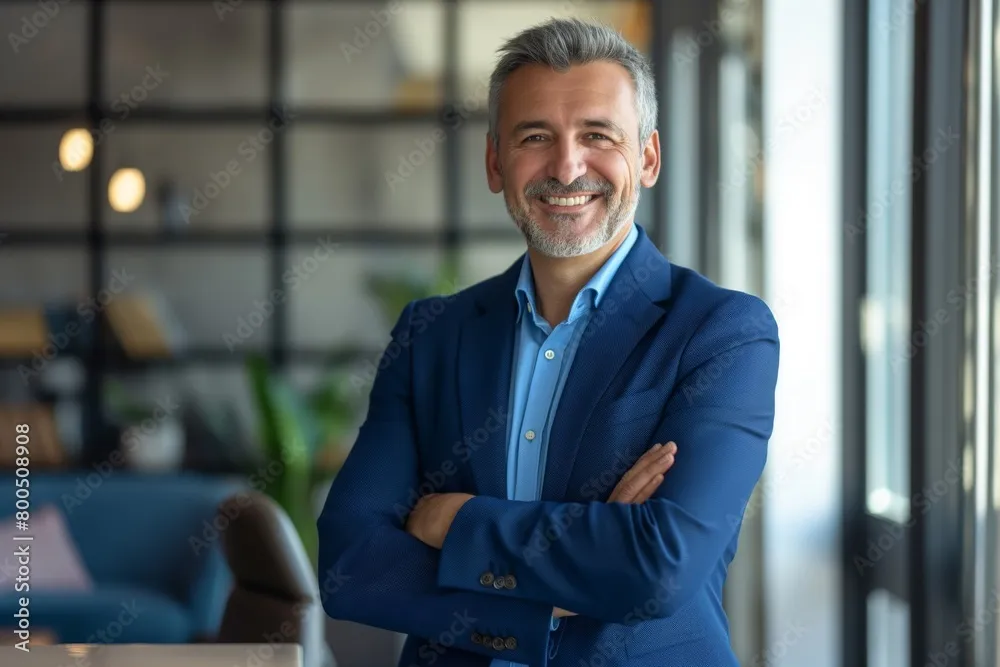 Happy middle aged business man ceo standing in office arms crossed. Smiling mature confident professional executive manager, proud lawyer, businessman leader wearing blue suit