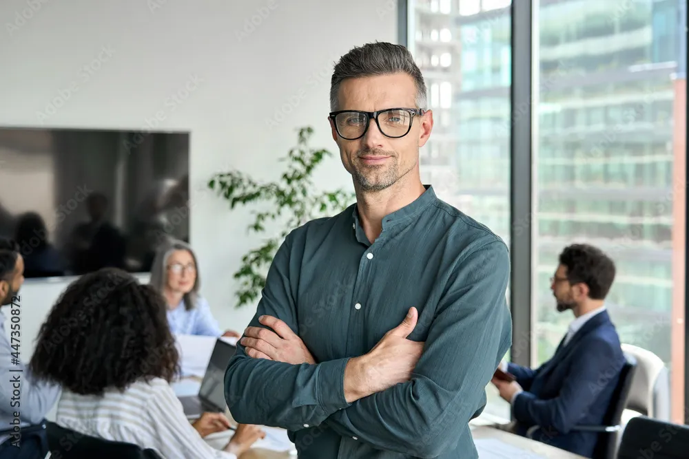 Smiling confident mature businessman leader looking at camera standing in office at team meeting. Male corporate leader ceo executive manager wearing glasses posing for business portrait arms folded.